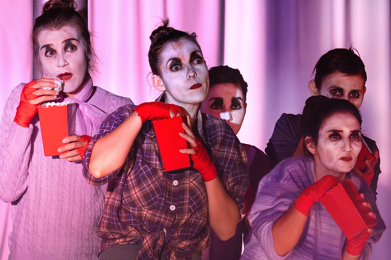 Cinco pessoas caracterizadas com maquilhagem de palco e luvas vermelhas observam atentamente enquanto comem pipocas, num cenário iluminado a rosa e roxo. atrizes de um projeto selecionado para apoio a espetáculos de teatro e dança pela Fundação GDA